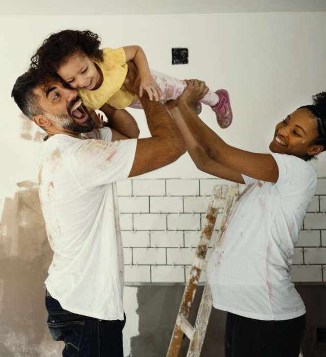 Un padre y una madre levantan de forma juguetona a su hija pequeña en una habitación que se está renovando.