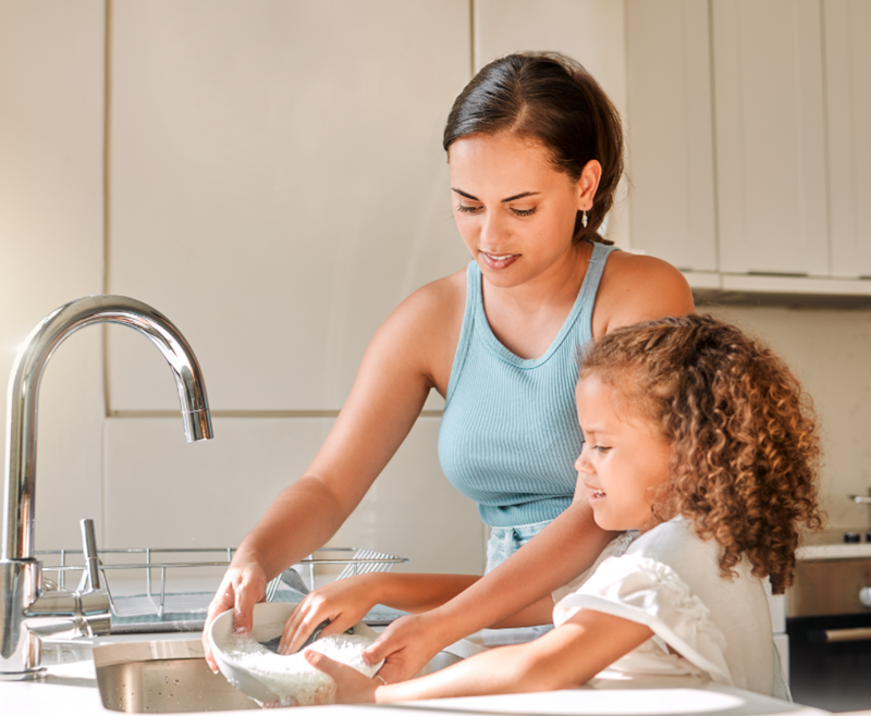 Una mujer de pie junto a un fregadero sostiene un plato mientras su hija pequeña usa un paño para lavarlo.