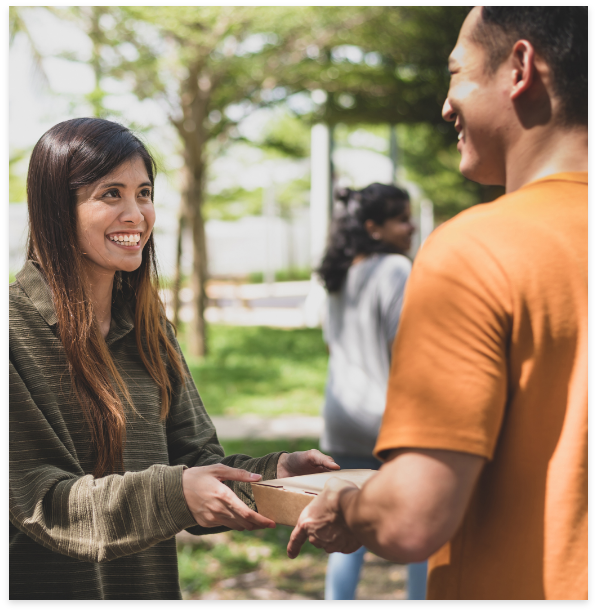 A woman hands a cardboard package to man in an outdoor park.