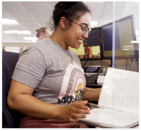 A woman sits at her workstation and flips through a binder of instructions.