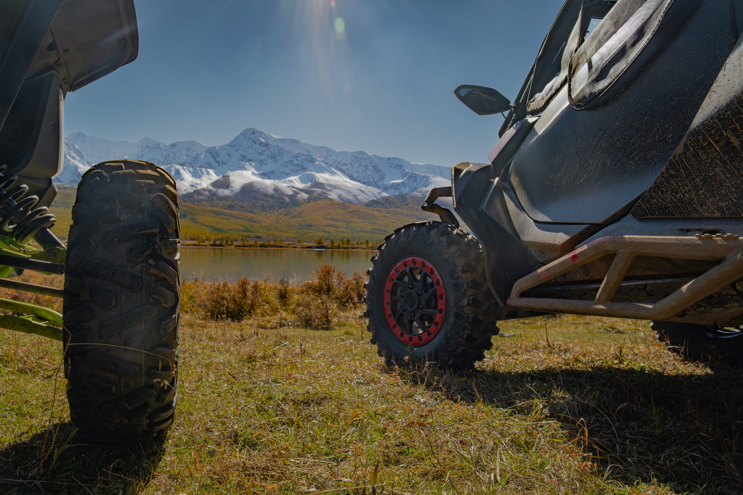 Close up of UTV off-road vehicles parked on a meadow by a mountain lake with snow peaks. Expedition, extreme adventure travel and overlanding concept.