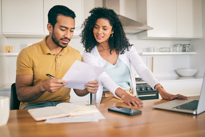 A couple sits at their kitchen table surrounded by papers and a laptop reviewing their finances.